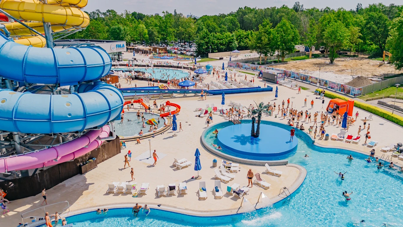 Kolorowe zjeżdżalnie wodne, liczne osoby korzystające z basenów i leżaków, wakacyjna atmosfera w otwartym aquaparku otoczonym zielenią.