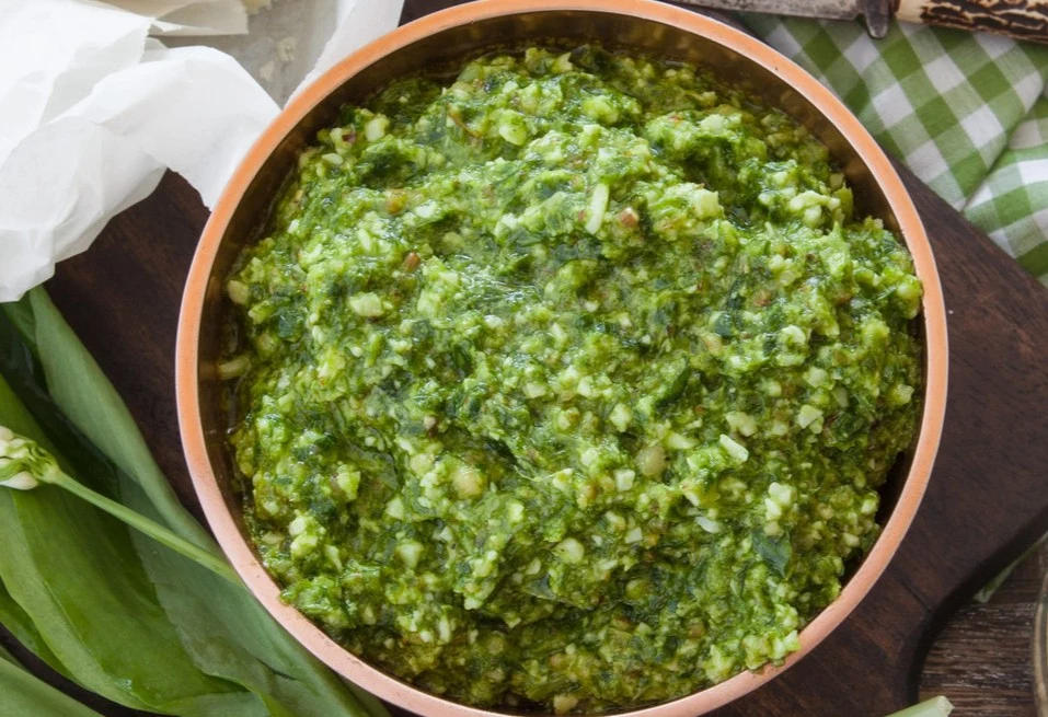 Pesto z młodych pędów czosnku to prawdziwy przysmak Miseczka pesto z młodych pędów czosnku z kawałkami orzechów na desce, obok liści czosnku niedźwiedziego.