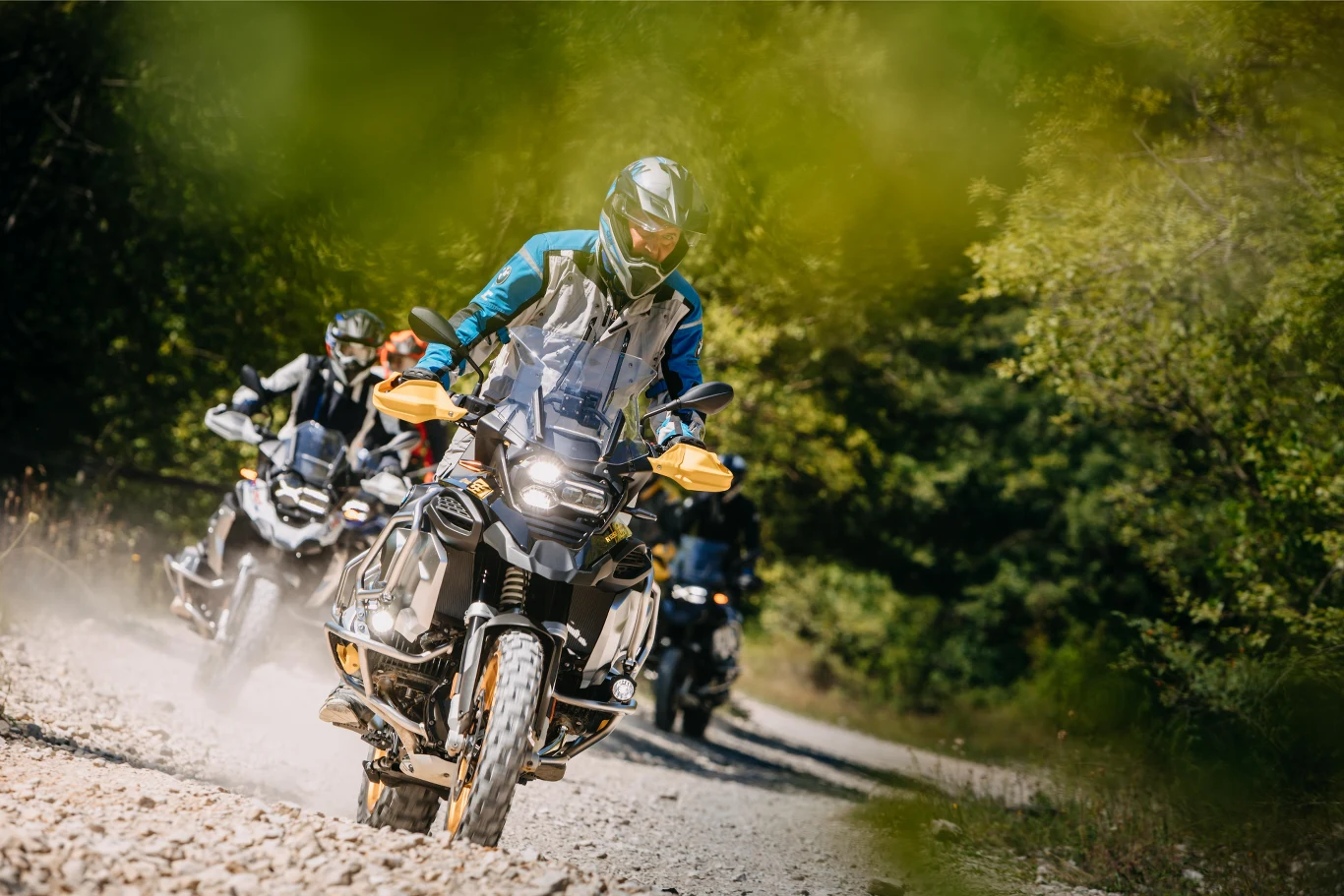 Grupa motocyklistów w kaskach i ochronnej odzieży jedzie po kamienistej leśnej drodze na nowoczesnych motocyklach terenowych, unosząc za sobą kurz.