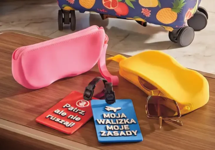 Kolorowe etui na okulary, przywieszki bagażowe z napisami „Patrz ale nie ruszaj!” i „Moja walizka moje zasady”, brązowe okulary przeciwsłoneczne oraz walizka w owocowy wzór w tle.
