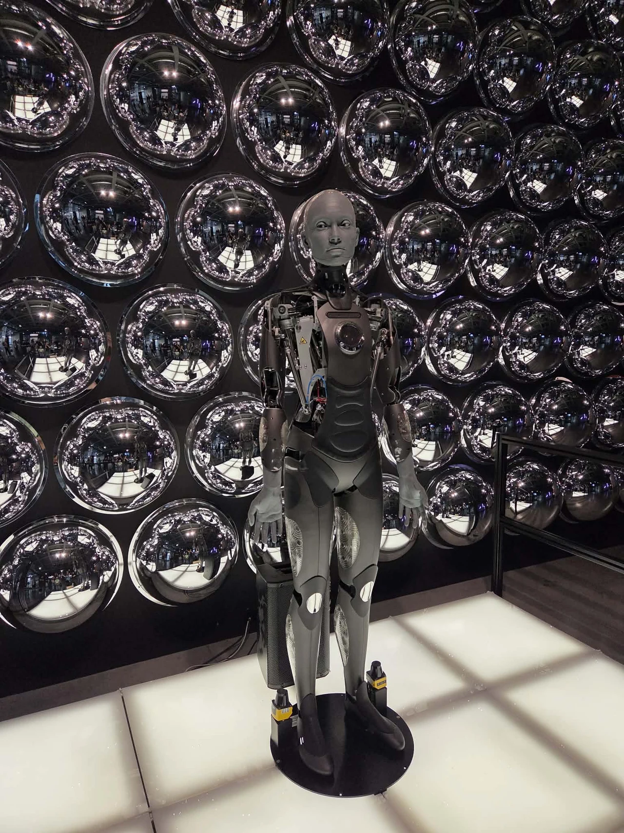 Humanoidalny robot o realistycznych rysach twarzy, stojący na jasnej podłodze, na tle ściany pokrytej błyszczącymi metalicznymi kulami.