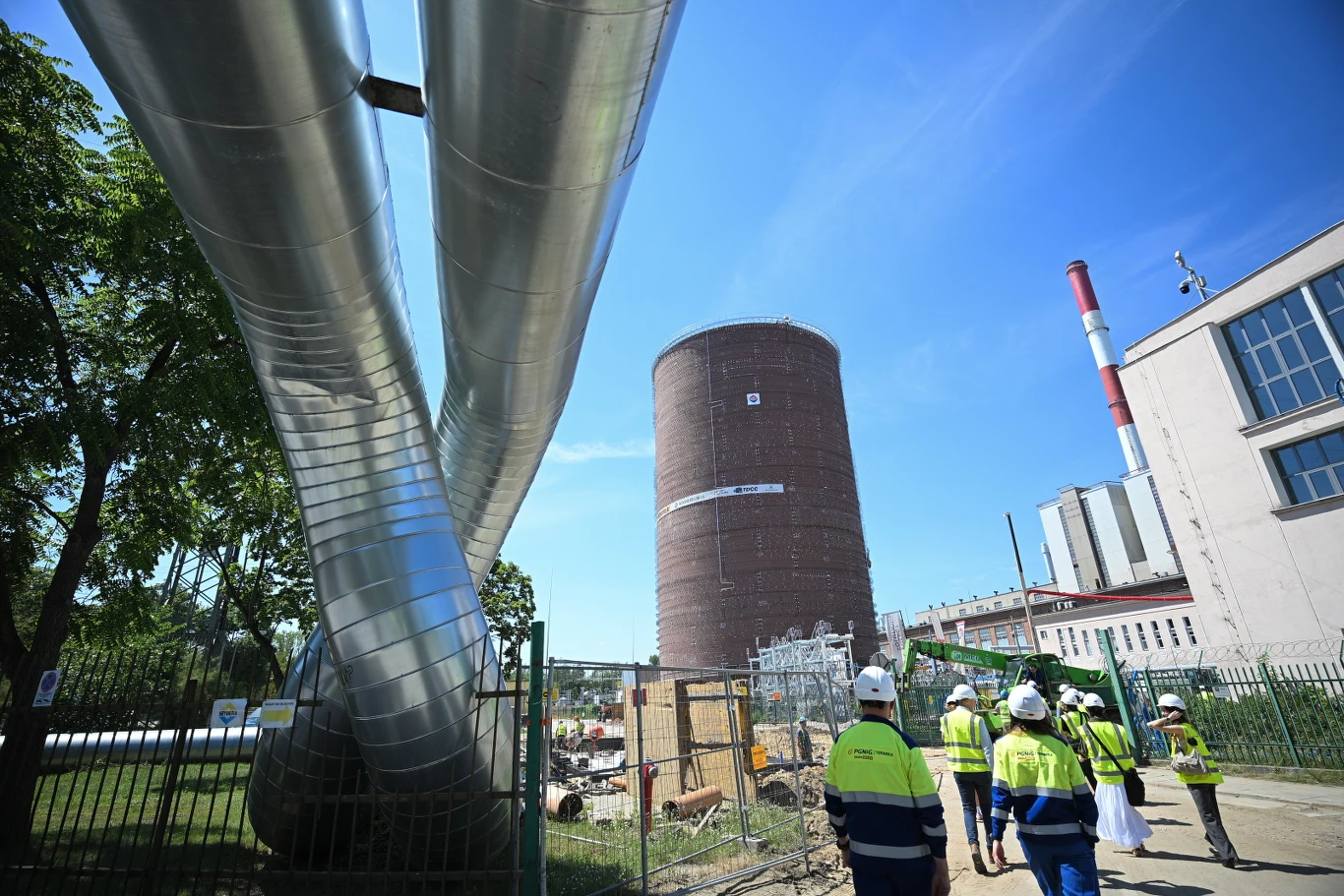 Grupa pracowników w odzieży ochronnej przemierza teren budowy przy dużych instalacjach rurowych i cylindrycznym zbiorniku, na tle obiektów przemysłowych oraz wysokiego komina.