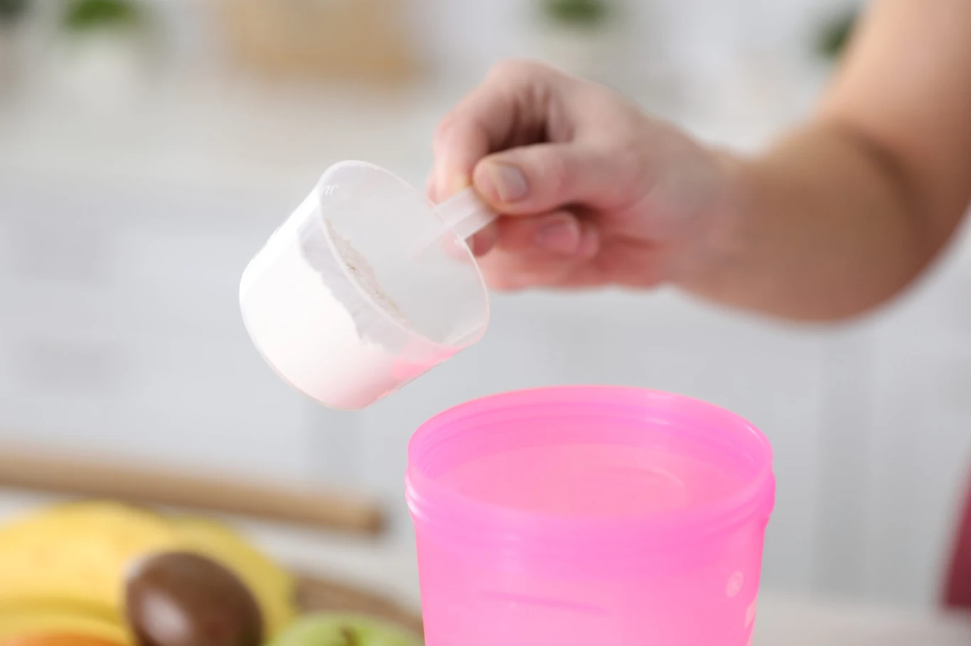 Ręka trzyma plastikową miarkę z białym proszkiem nad różowym shakerem.