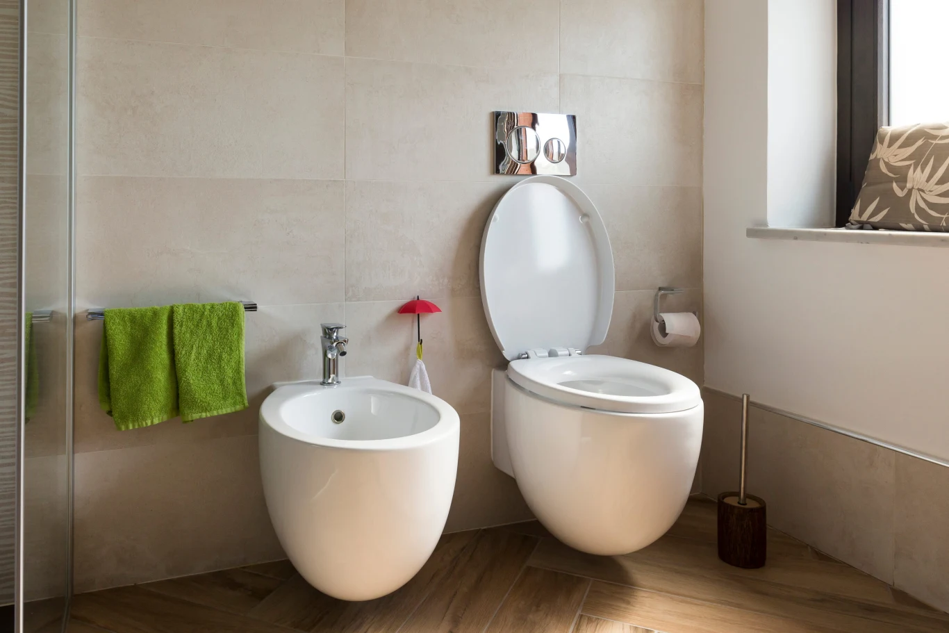 Bidet to coraz popularniejsze urządzenie w polskich łazienkach, ale wymaga osobnego odpływu kanalizacyjnego. Nowoczesna łazienka z podwieszaną toaletą, bidetem, zielonym ręcznikiem i oknem z poduszką.