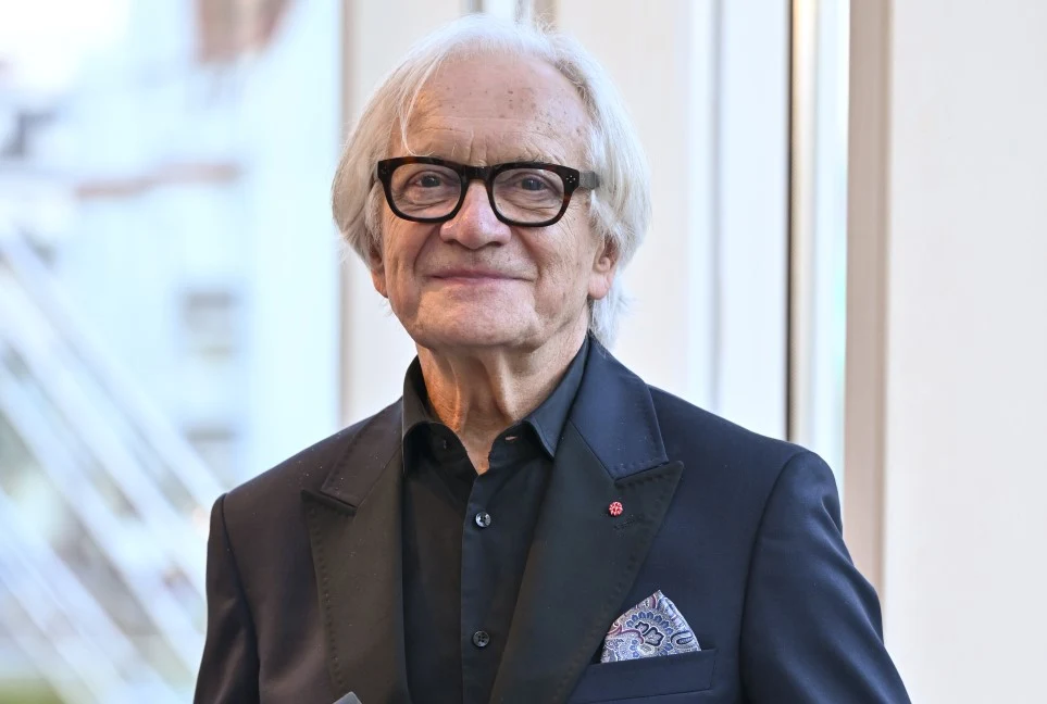 Andrzej Seweryn na gali Festiwalu Dwa Teatry, 19 czerwca 2026 Starszy mężczyzna z siwymi włosami i okularami w eleganckim, ciemnym garniturze, ozdobionym poszetką, stoi przy dużym oknie, uśmiechając się lekko.