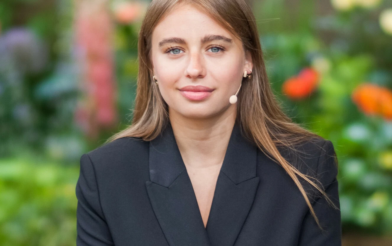 Oliwia Bieniuk Oliwia Bieniuk w czarnej marynarce i z mikrofonem na uchu na tle roślinności.