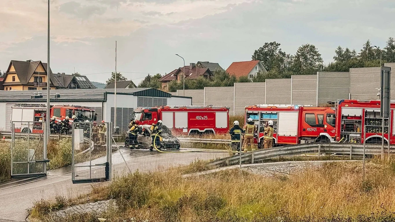 Strażacy gaszący samochód przy wsparciu kilku wozów strażackich, otoczeni barierkami ochronnymi na terenie drogi w pobliżu zabudowań mieszkalnych.