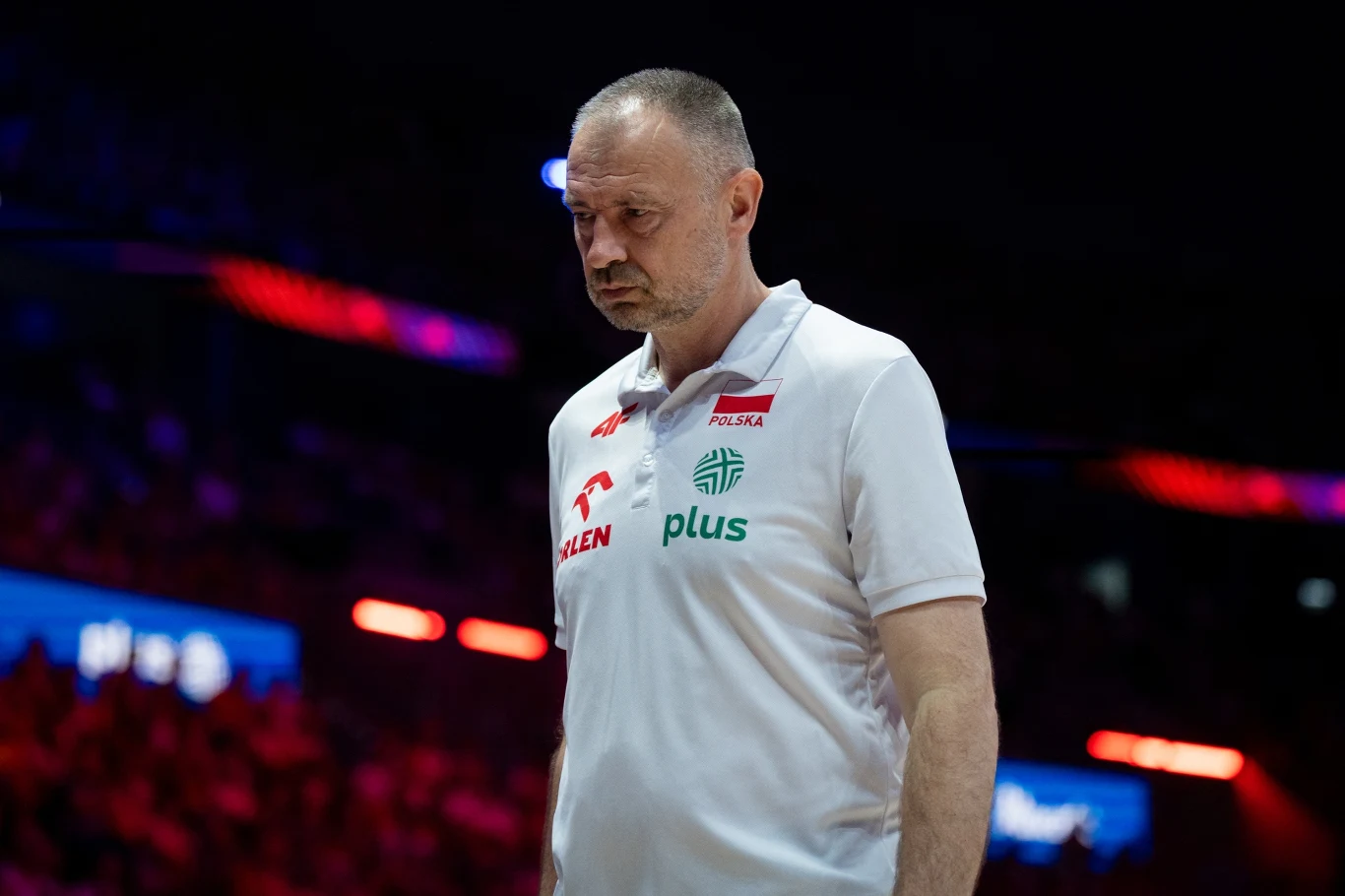 Mężczyzna w białej koszulce z logo Orlen i Plus oraz napisem Polska, stoi w skupieniu na tle rozświetlonej, sportowej hali.