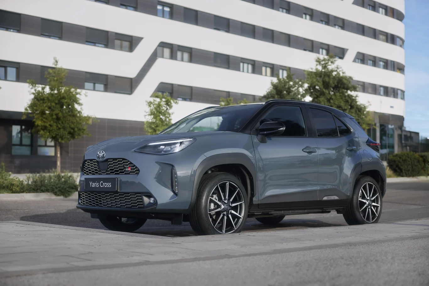 Szary crossover Toyota Yaris Cross na dużych, stylowych felgach, zaparkowany na tle nowoczesnego, wielopiętrowego budynku.