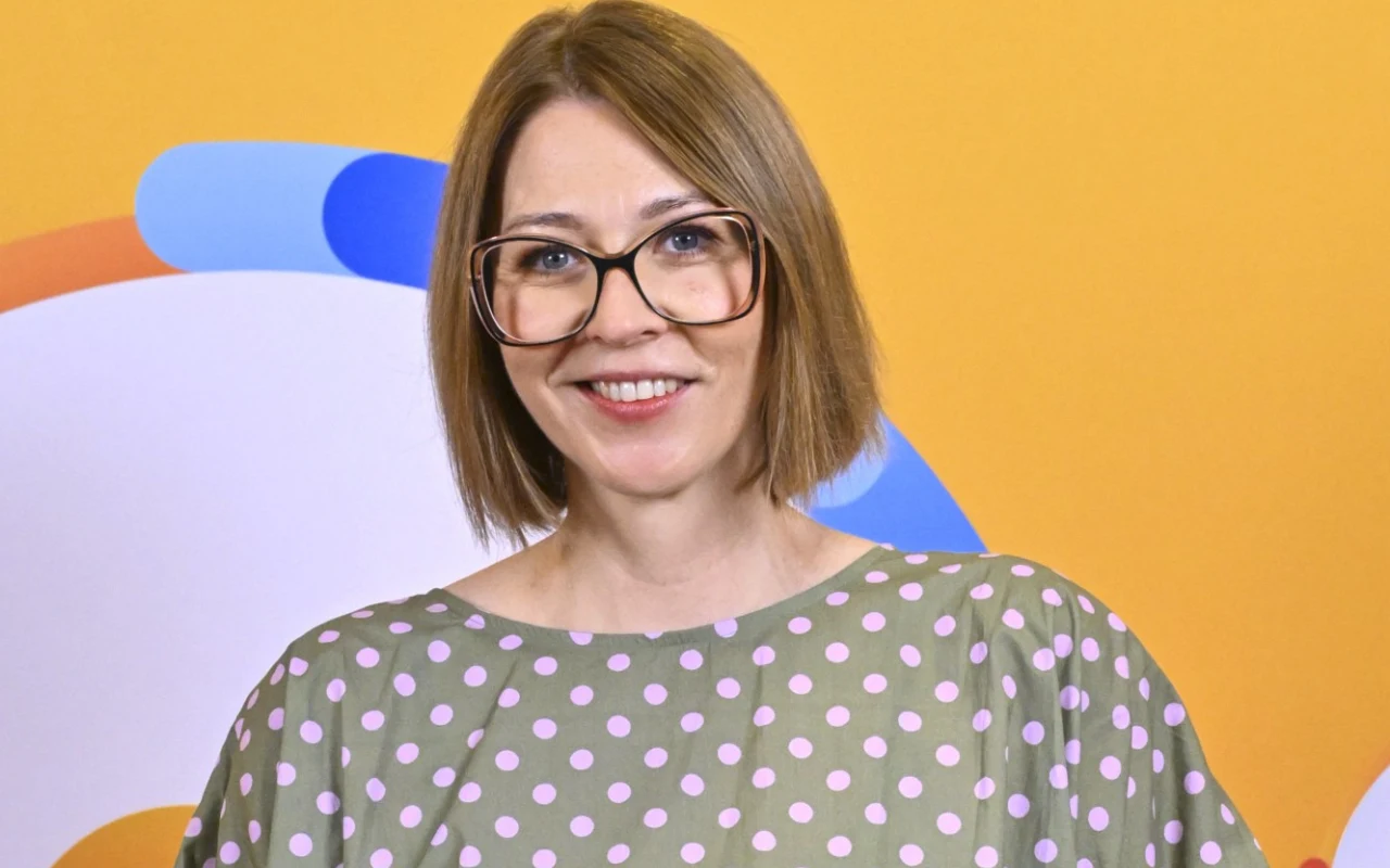 Marta Eljasiak-Dziki Kobieta z krótkimi, prostymi włosami w dużych okularach, ubrana w szarą sukienkę w różowe grochy, stoi na tle żółtej ściany z kolorowymi, abstrakcyjnymi kształtami.