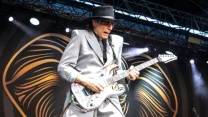 Muzyk w jasnoszarym garniturze i kapeluszu grający na gitarze elektrycznej na dużej scenie z charakterystyczną grafiką w tle.