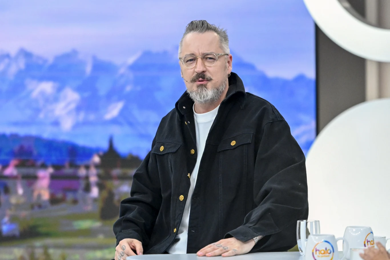 Piotr Kędzierski w "halo tu polsat" Mężczyzna z siwą brodą i okularami w czarnej kurtce stoi przy stole w nowoczesnym studiu telewizyjnym, na tle rozmytego widoku gór.