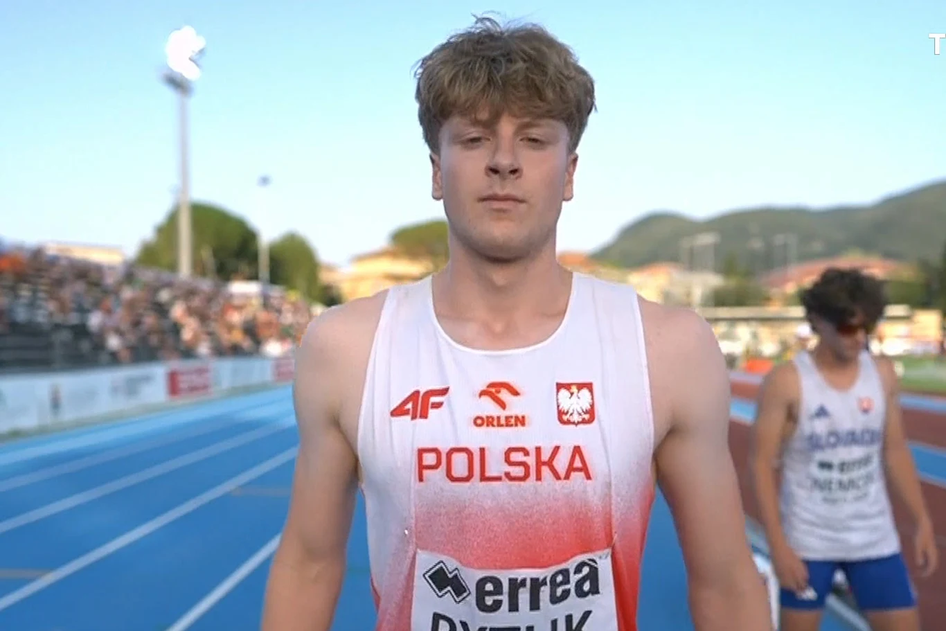 Lekkoatleta w stroju reprezentacji Polski stoi na bieżni stadionu lekkoatletycznego, w tle widoczni inni zawodnicy oraz trybuny z kibicami.