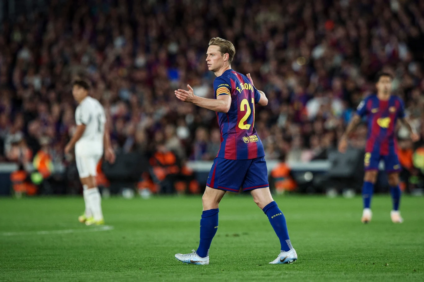 Piłkarz w stroju FC Barcelony z numerem 21 na plecach gestykuluje w trakcie meczu, w tle widoczni są inni zawodnicy oraz zapełnione trybuny stadionu.