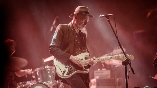 Gitarzysta w kapeluszu grający na scenie elektryczną gitarę, w tle widoczna perkusja i sceniczne oświetlenie.