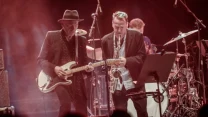 Zespół muzyczny podczas występu na scenie, gitarzysta w kapeluszu gra na gitarze elektrycznej obok saksofonisty, w tle widoczny perkusista przy zestawie perkusyjnym.