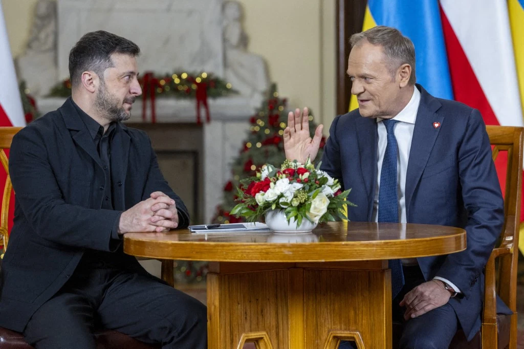 Dwóch mężczyzn siedzi naprzeciw siebie przy okrągłym drewnianym stole udekorowanym bukietem kwiatów, prowadząc rozmowę; w tle widoczne są flagi i kompozycje świąteczne.