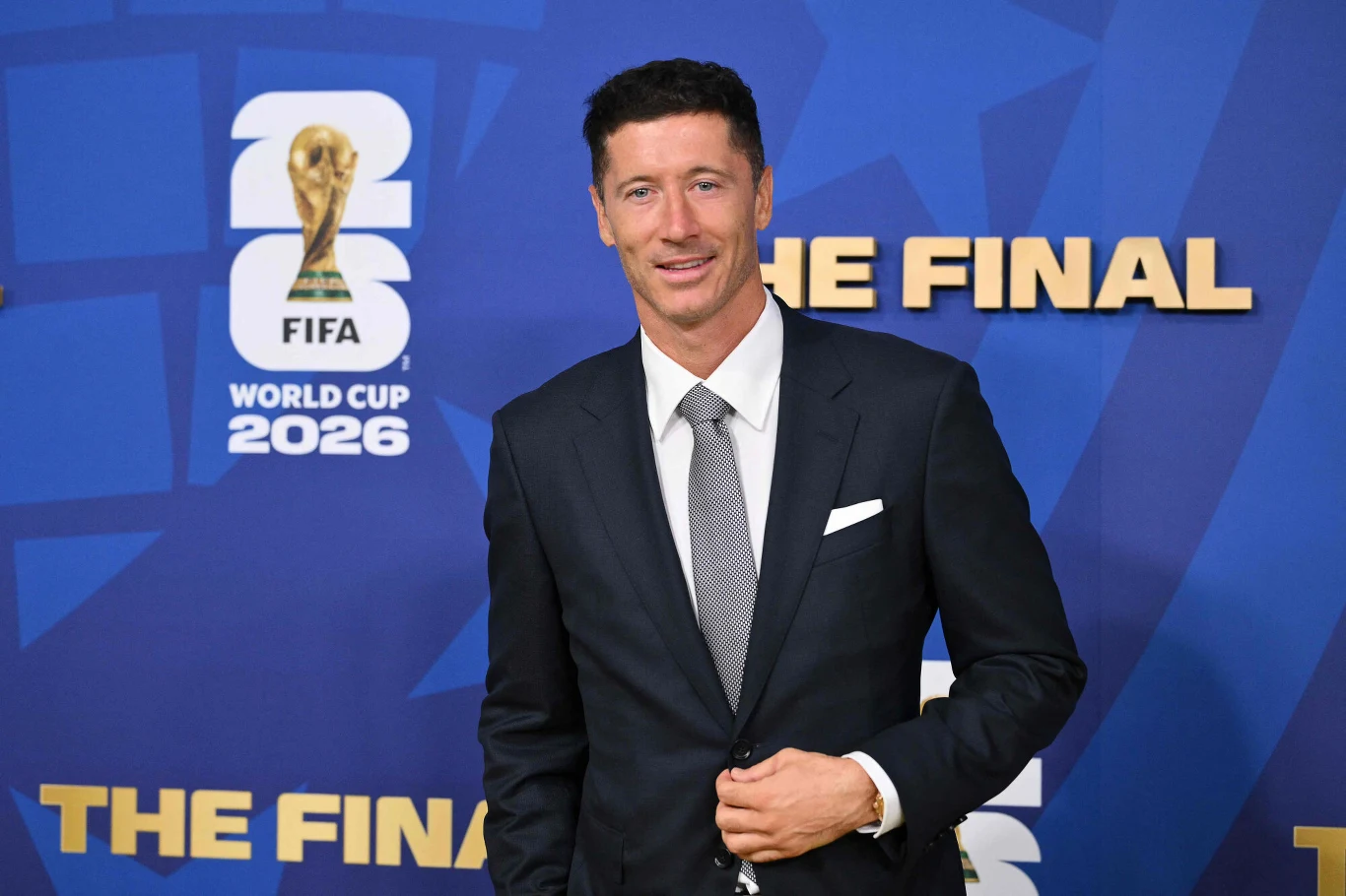 Elegancko ubrany mężczyzna w ciemnym garniturze i krawacie stoi na tle ściany z logo Mistrzostw Świata FIFA 2026.