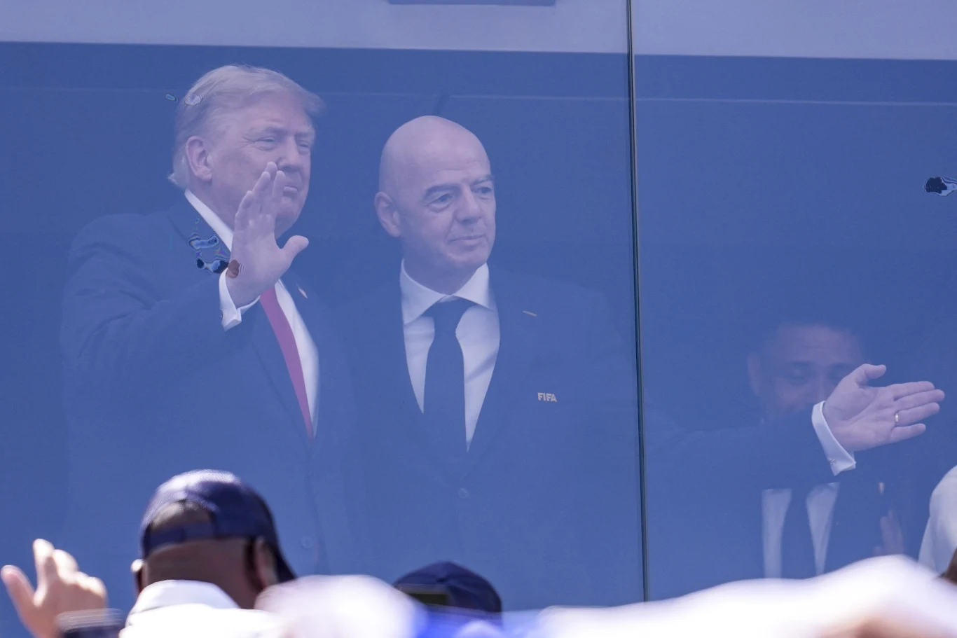 Donald Trump i Gianni Infantino Donald Trump i Gianni Infantino stoją w garniturach za szybą i machają do tłumu na stadionie.