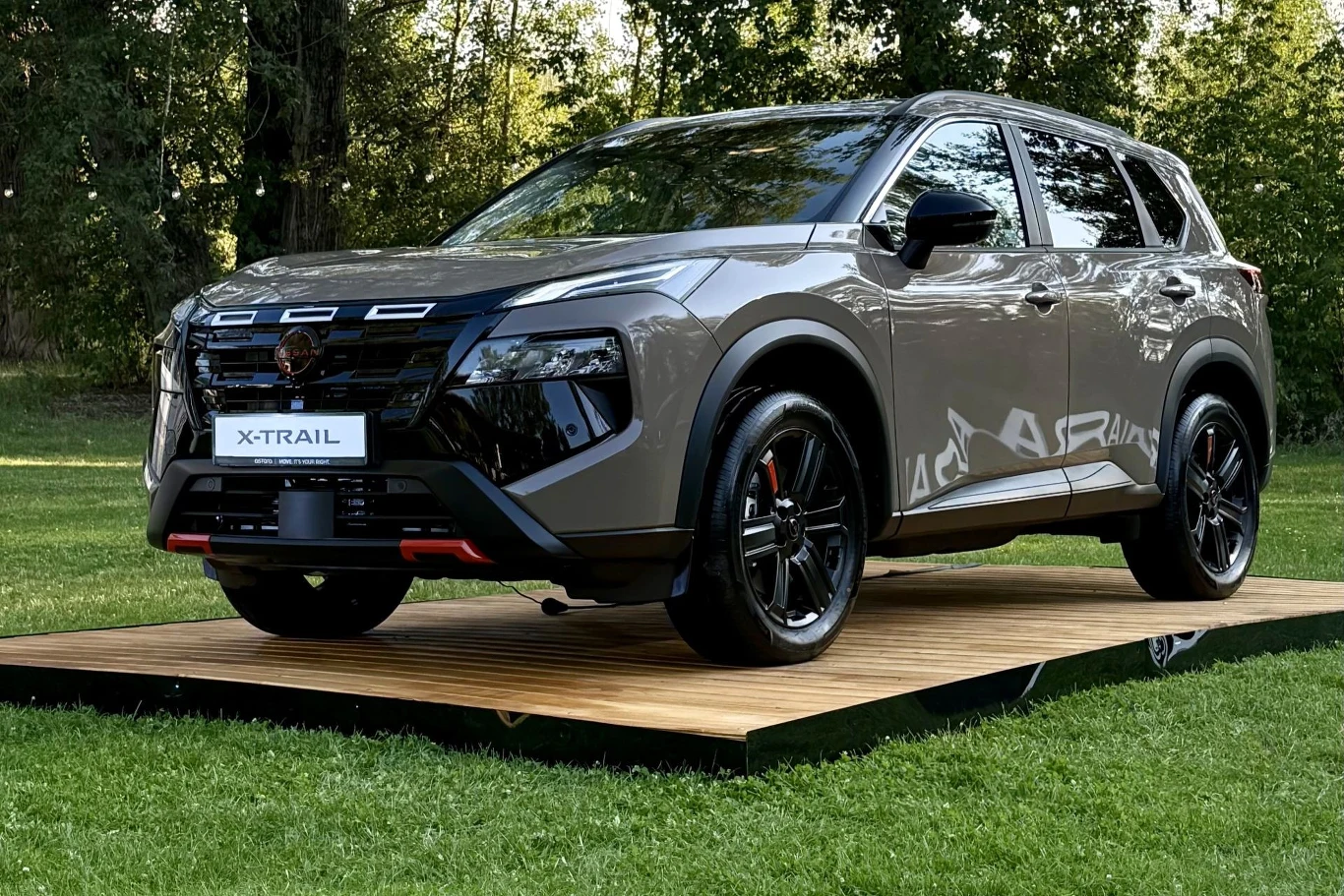 Ciemnoszary SUV marki Nissan X-Trail zaprezentowany na drewnianym podwyższeniu, na tle zielonego trawnika i drzew.