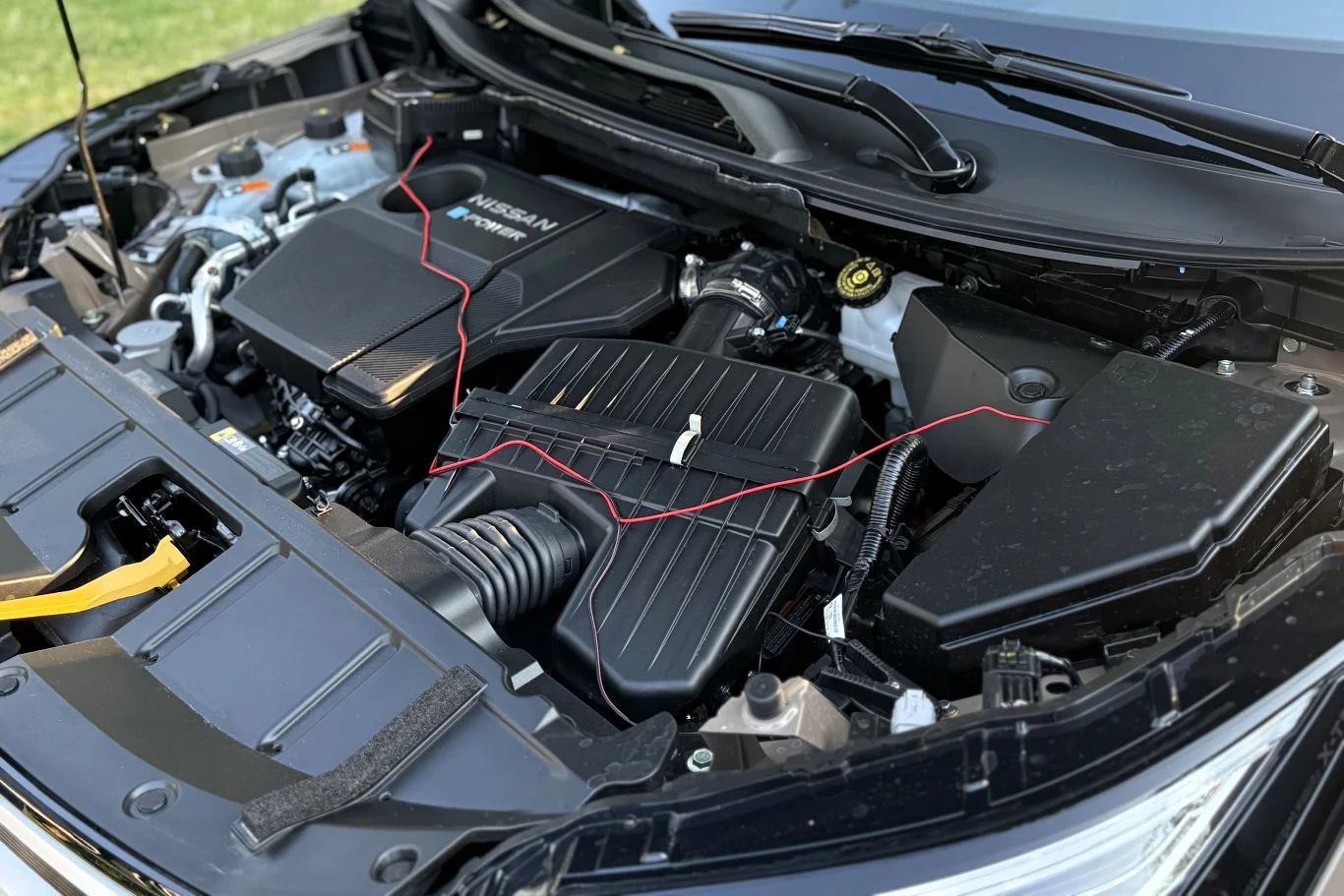 Silnik samochodu marki Nissan z widocznymi elementami układu dolotowego, osłoną silnika oraz przewodami elektrycznymi rozłożonymi na podzespołach.