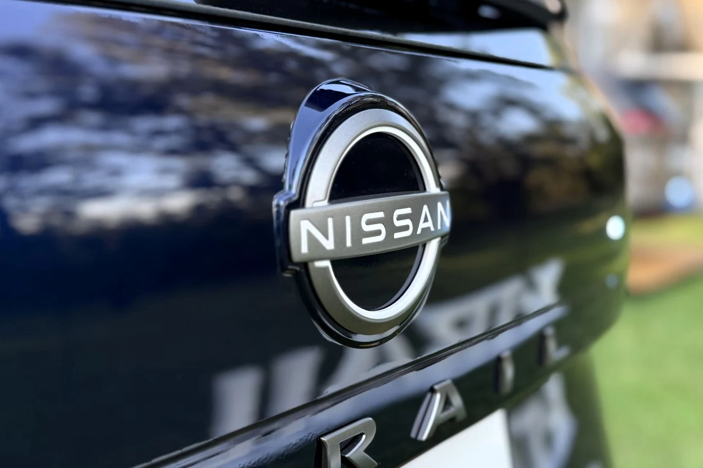 Duże, srebrne logo marki Nissan umieszczone centralnie na błyszczącej, ciemnej klapie bagażnika samochodu.