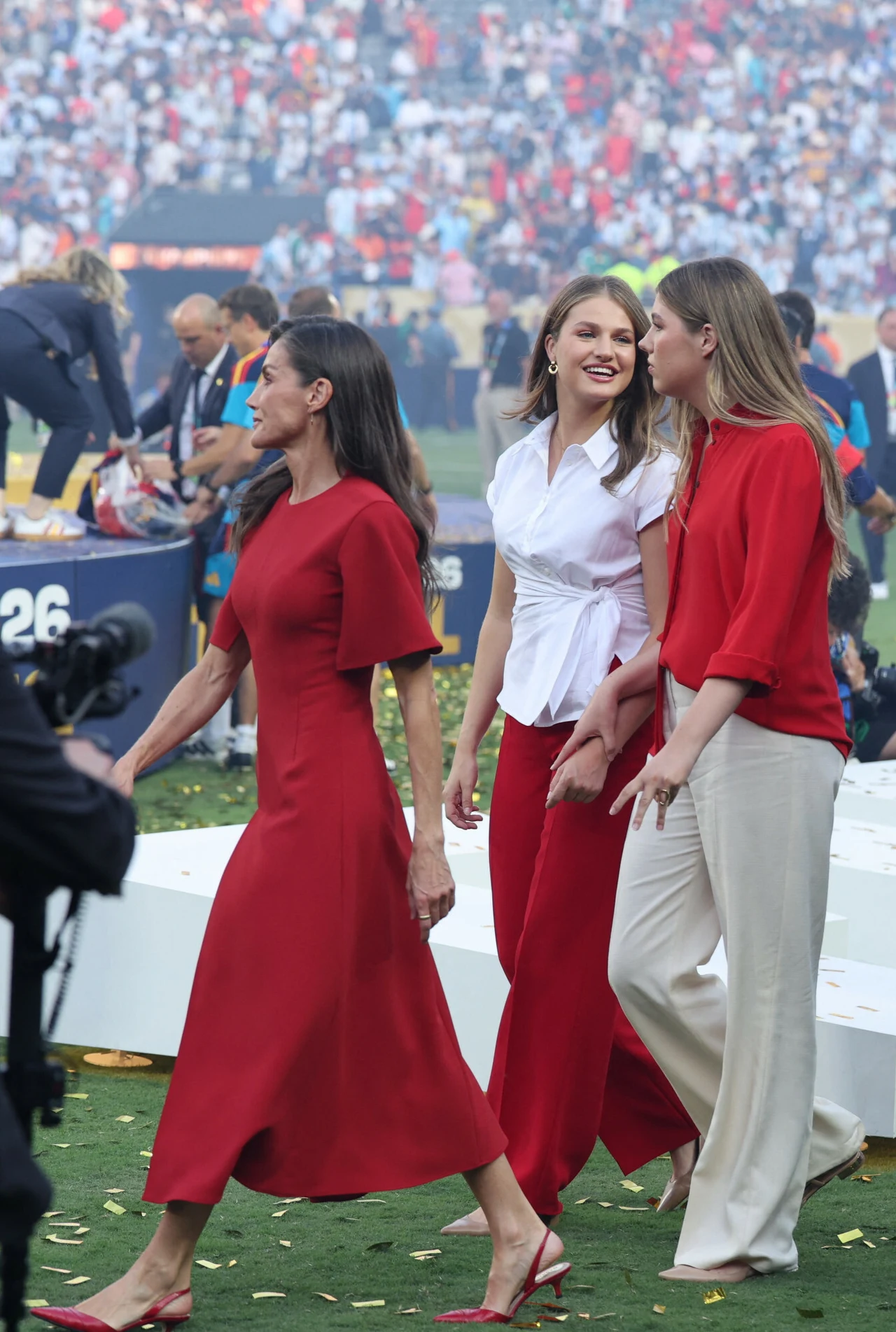 Trzy elegancko ubrane kobiety idące po murawie stadionu, w otoczeniu konfetti, tłumu kibiców oraz telewizyjnych kamer, ubrane w czerwone i białe stylizacje.
