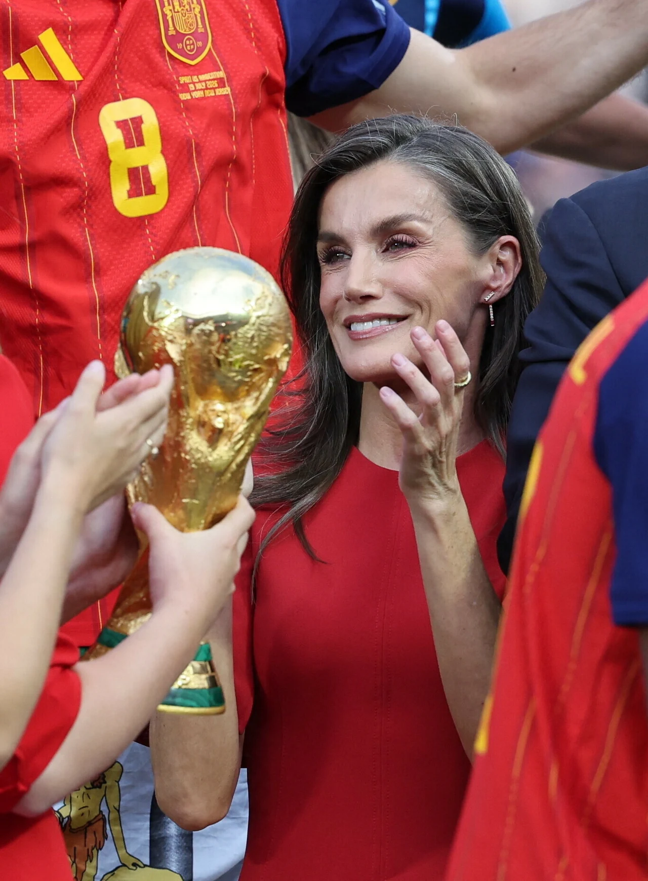 Kobieta w czerwonej sukience z uśmiechem spogląda na puchar FIFA World Cup trzymany przez osobę w koszulce reprezentacji Hiszpanii.