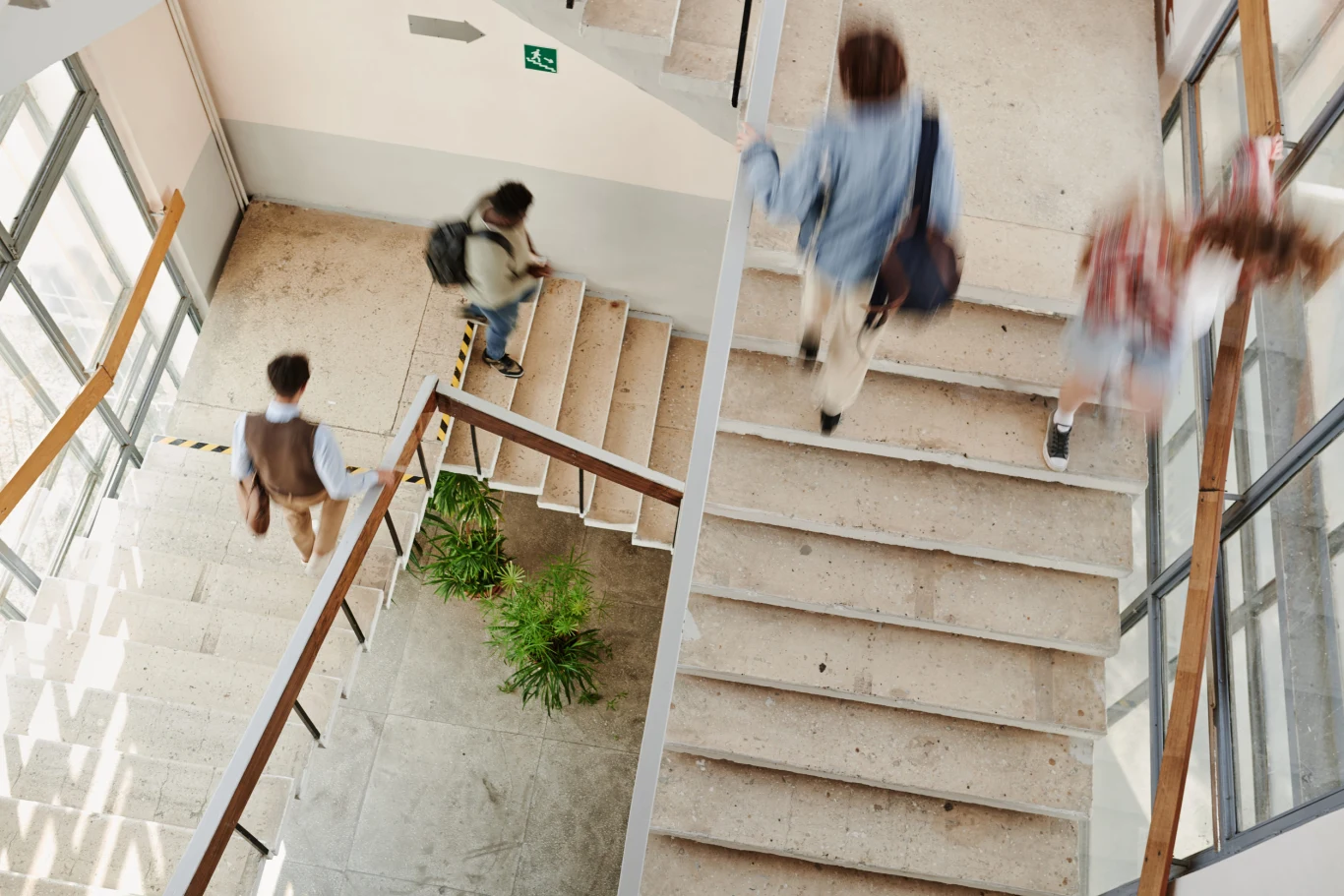 Uniki na półpiętrze. Dlaczego boimy się własnych sąsiadów? Kilka osób z plecakami schodzi po szerokich schodach w jasnym budynku, w pobliżu których ustawiono zielone rośliny.