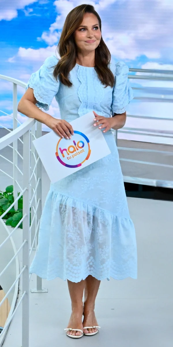 Paulina Sukut-Jeżyna wyglądała świetnie w modnej, koronkowej sukience Kobieta w jasnoniebieskiej sukience z falbanami trzyma plan programu z kolorowym logo, stoi przy białej balustradzie na tle słonecznego nieba.