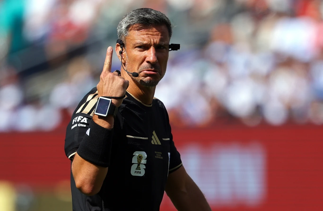 Sędzia piłkarski w czarnej koszulce z emblematem FIFA, unoszący dwa palce w górę i wyposażony w zestaw słuchawkowy oraz smartwatch na ręce.