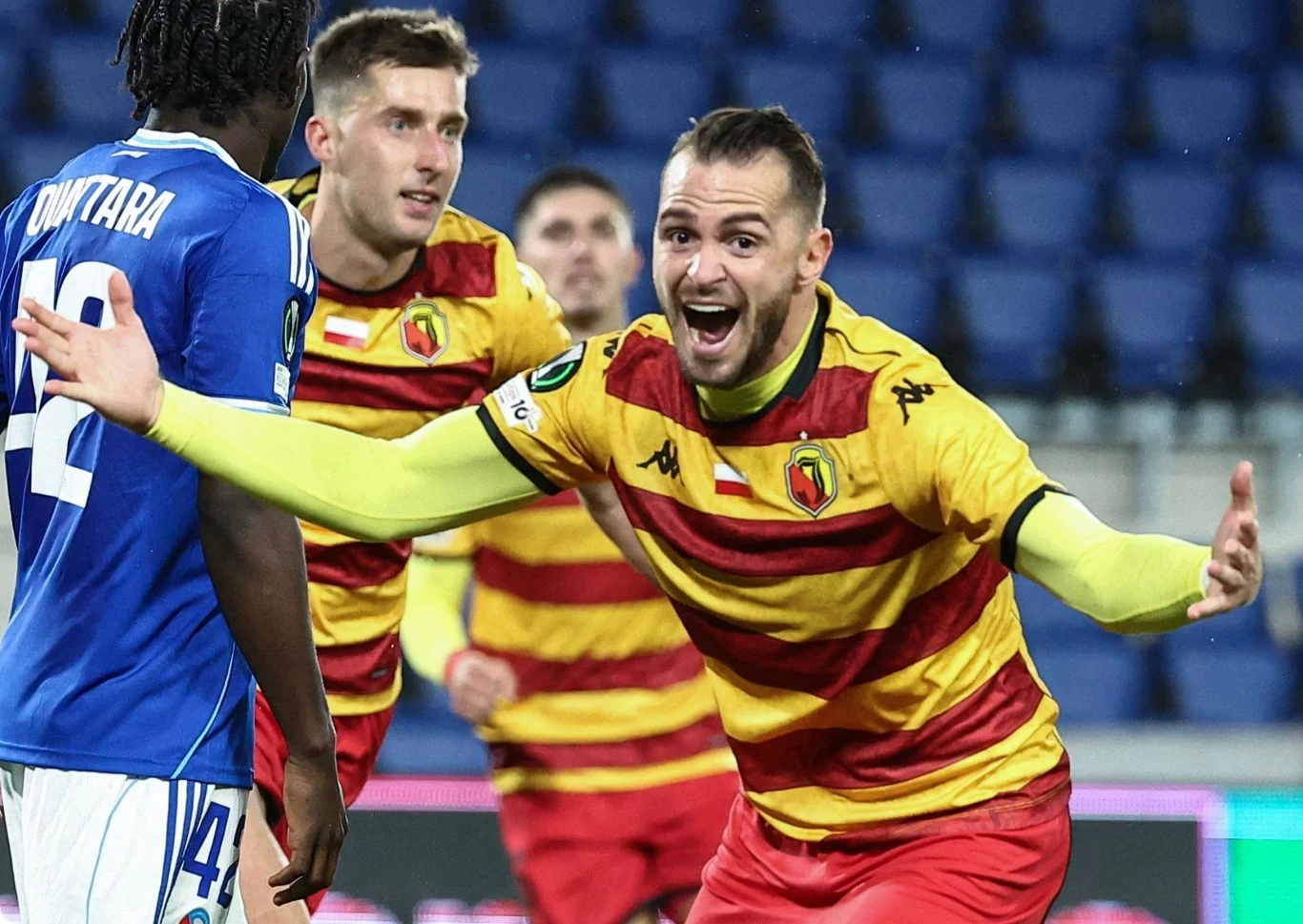 Jesus Imaz Jagiellonia Białystok Piłkarz w żółto-czerwonej koszulce z uniesionymi rękami w geście radości podczas meczu, z innymi zawodnikami i pustymi trybunami w tle.