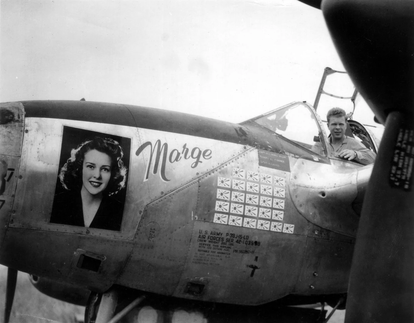 Amerykański myśliwiec z II wojny światowej z portretem kobiety i napisem „Marge” na kadłubie oraz licznymi symbolami zwycięstw, w kokpicie pilot w wojskowym kombinezonie.