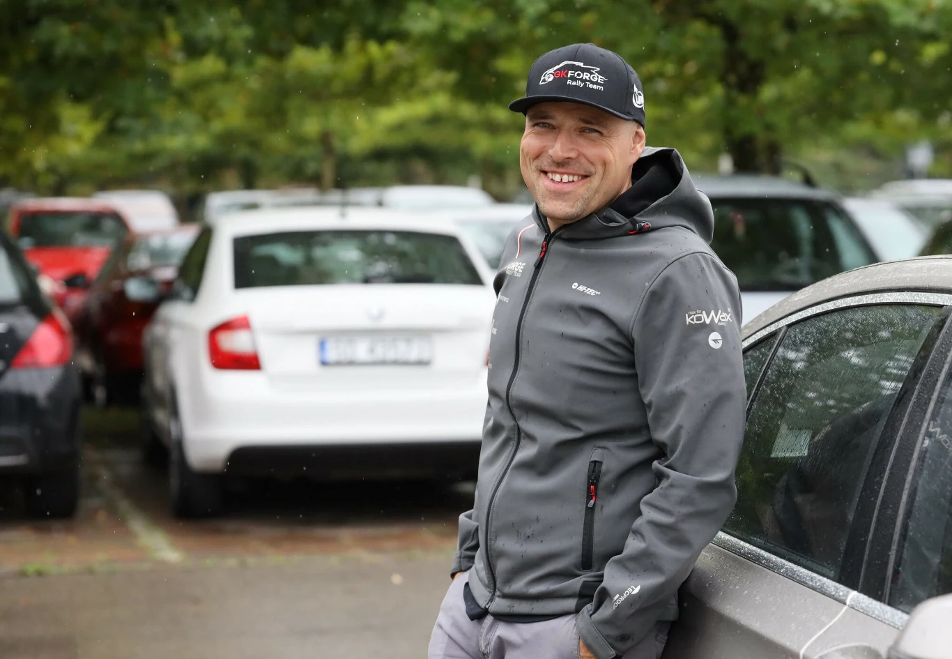 Mężczyzna w sportowej kurtce i czapce z daszkiem oparty o samochód, w tle zaparkowane auta i drzewa.