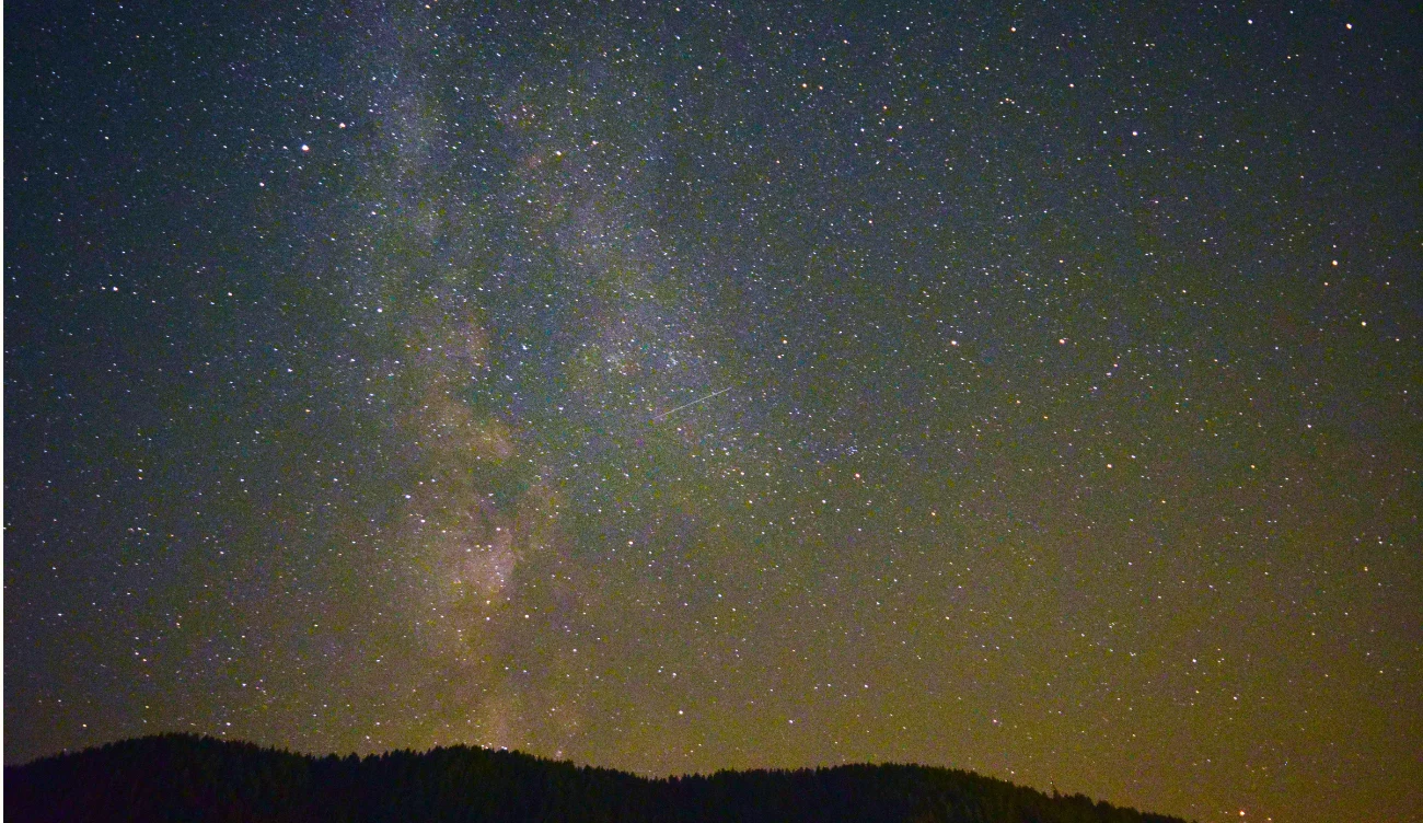 O której godzinie najlepiej oglądać rój meteorów Perseidy? Rozświetlony pas Drogi Mlecznej na nocnym niebie nad ciemnym zarysem gór podczas roju meteorów Perseidów.