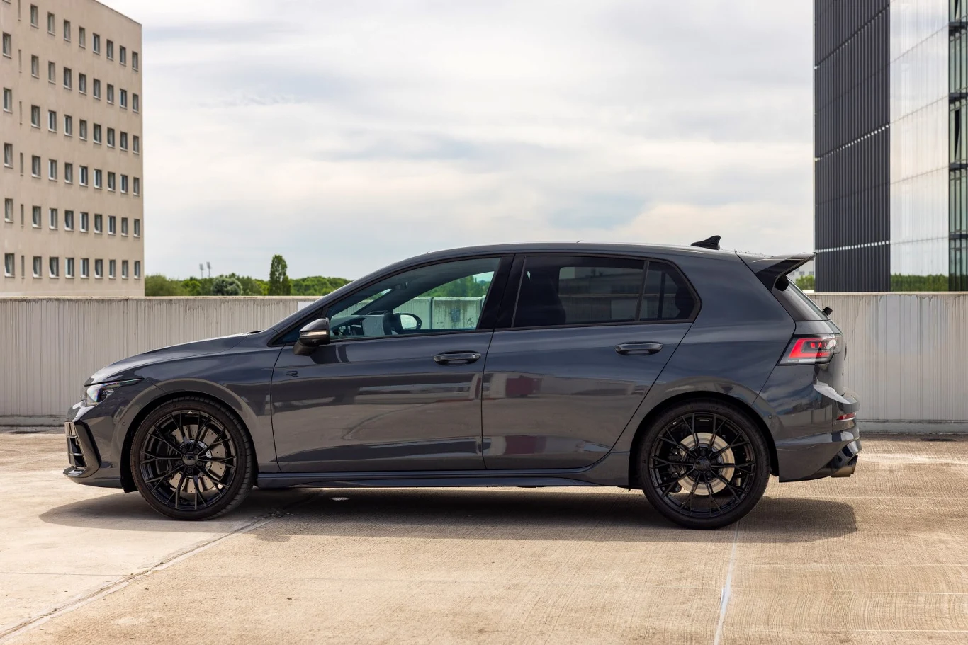 Szary, nowoczesny hatchback z przyciemnianymi szybami i czarnymi felgami stoi zaparkowany na betonowym parkingu w otoczeniu wysokich budynków.