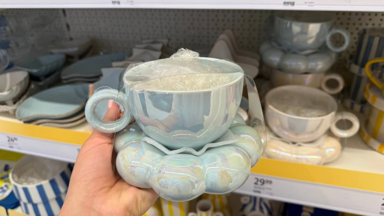 Jasnoniebieska filiżanka z połyskiem ustawiona na miękkiej, puszystej podstawce w tym samym kolorze, trzymana w dłoni, na tle innych porcelanowych naczyń na sklepowej półce.