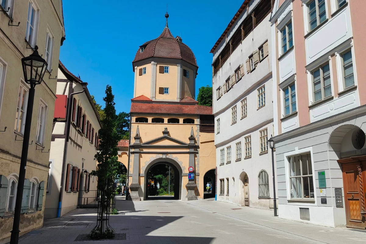 Zabytkowa brama miejska z wieżą o ośmiokątnym dachu, otoczona dobrze zachowanymi kamienicami i jasną, szeroką ulicą w słoneczny dzień.