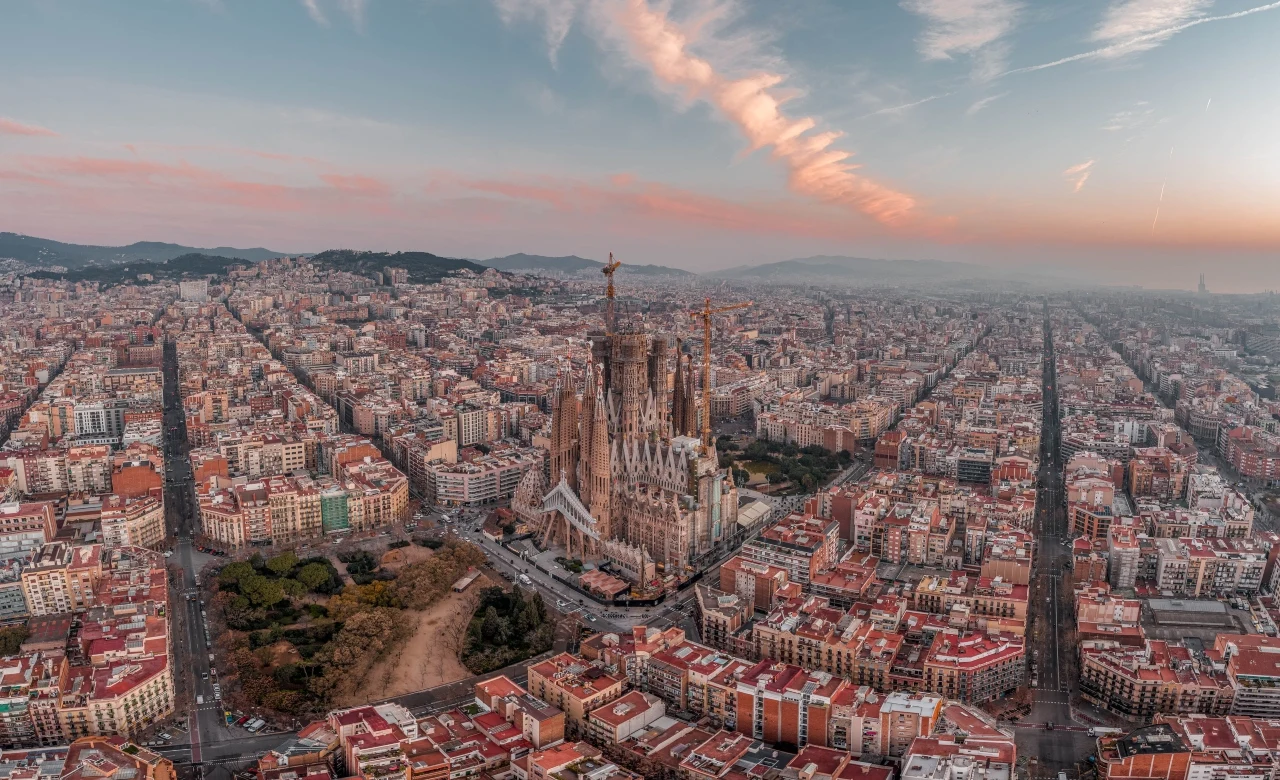 Najwyższa świątynia Europy - Sagrada Família w Barcelonie Sagrada Familia górująca nad symetryczną siatką ulic Barcelony, otoczona gęstą zabudową miasta o poranku.