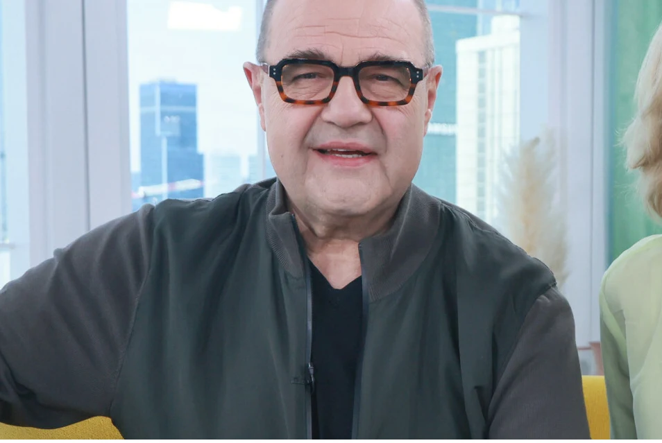 Cezary Żak w okularach o grubych oprawkach, ubrany w ciemną koszulkę i kurtkę, siedzi na żółtej sofie na tle dużych okien z widokiem na nowoczesne budynki.
