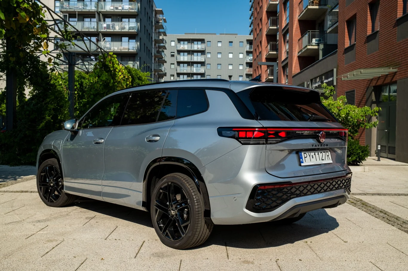 Srebrny SUV z czarnymi felgami zaparkowany na tle nowoczesnych budynków mieszkalnych, otoczony zielenią i miejską infrastrukturą.