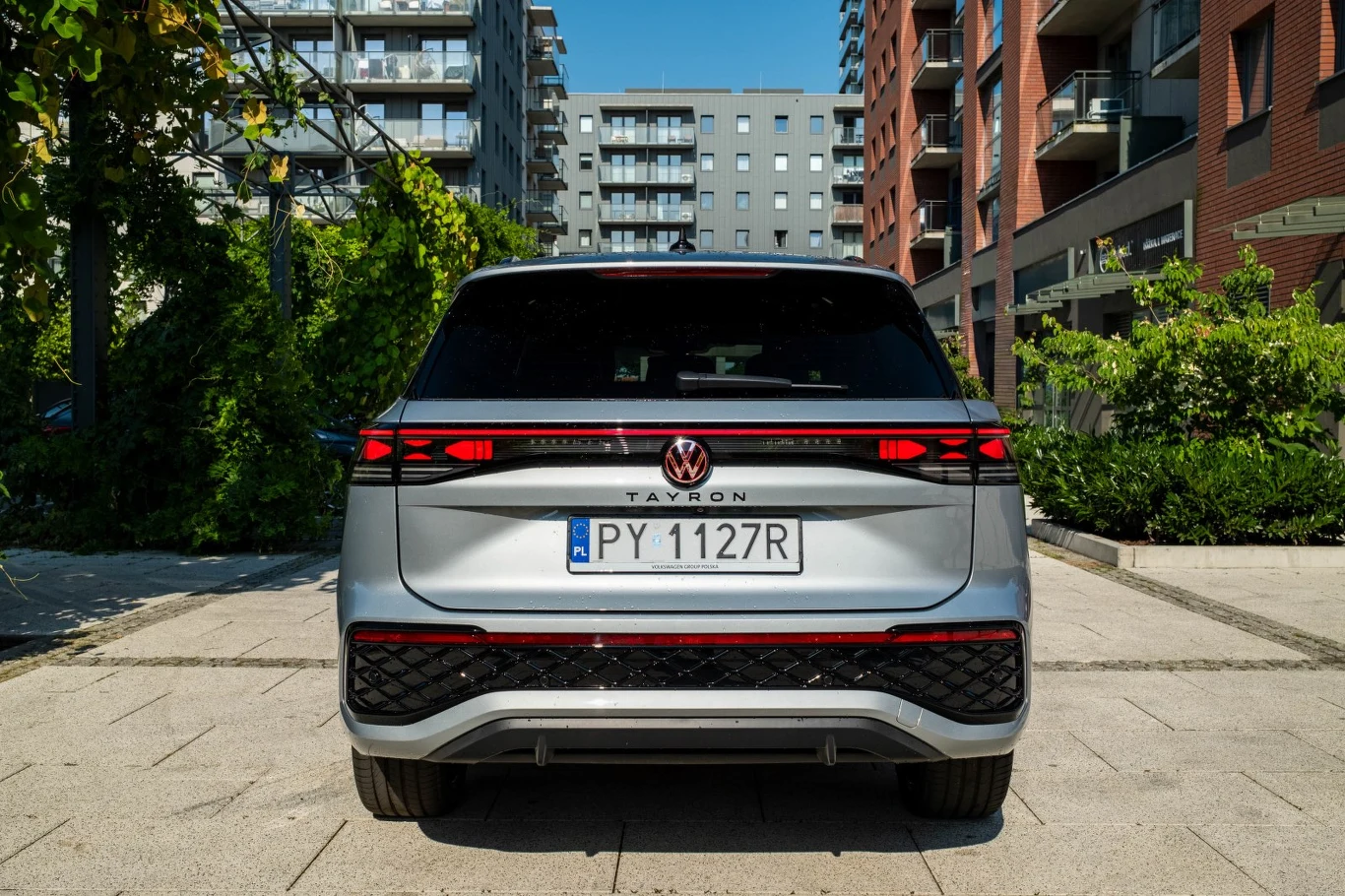 Srebrny Volkswagen Tayron zaparkowany na tle nowoczesnych budynków mieszkalnych, widoczny z tyłu z dobrze wyeksponowanymi tylnymi lampami LED i charakterystyczną tablicą rejestracyjną.