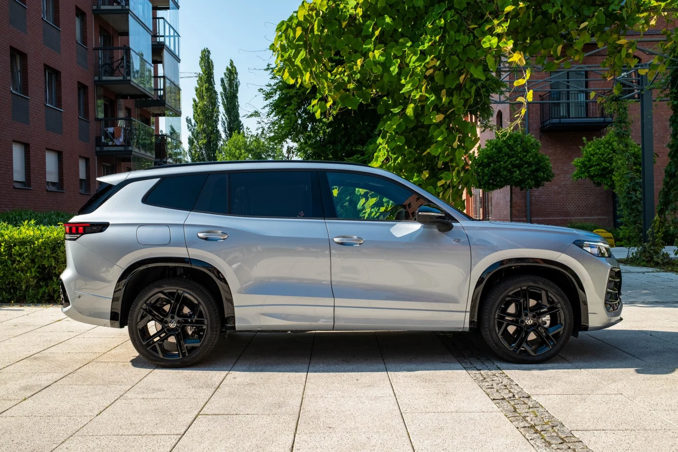 Srebrny SUV o nowoczesnej sylwetce zaparkowany na szerokim chodniku w otoczeniu miejskich budynków i zielonych drzew.