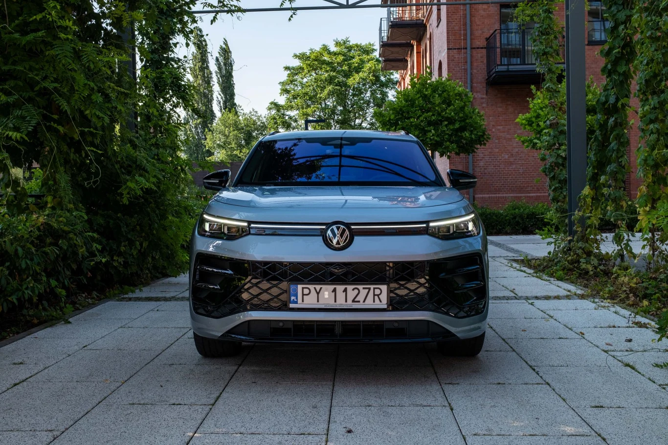 Srebrny Volkswagen zaparkowany na szerokiej, wyłożonej płytami alejce, otoczony zielenią i ceglaną zabudową mieszkalną po prawej stronie.