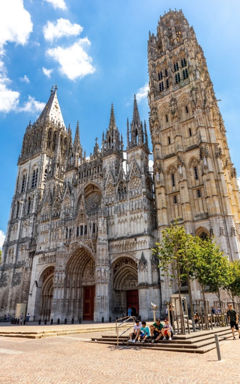 4. miejsce - katedra Najświętszej Marii Panny w Rouen Gotycka katedra z bogato zdobioną fasadą, strzelistymi wieżami i licznymi detalami architektonicznymi, przed budynkiem kilku siedzących na ławce ludzi oraz młode drzewa.