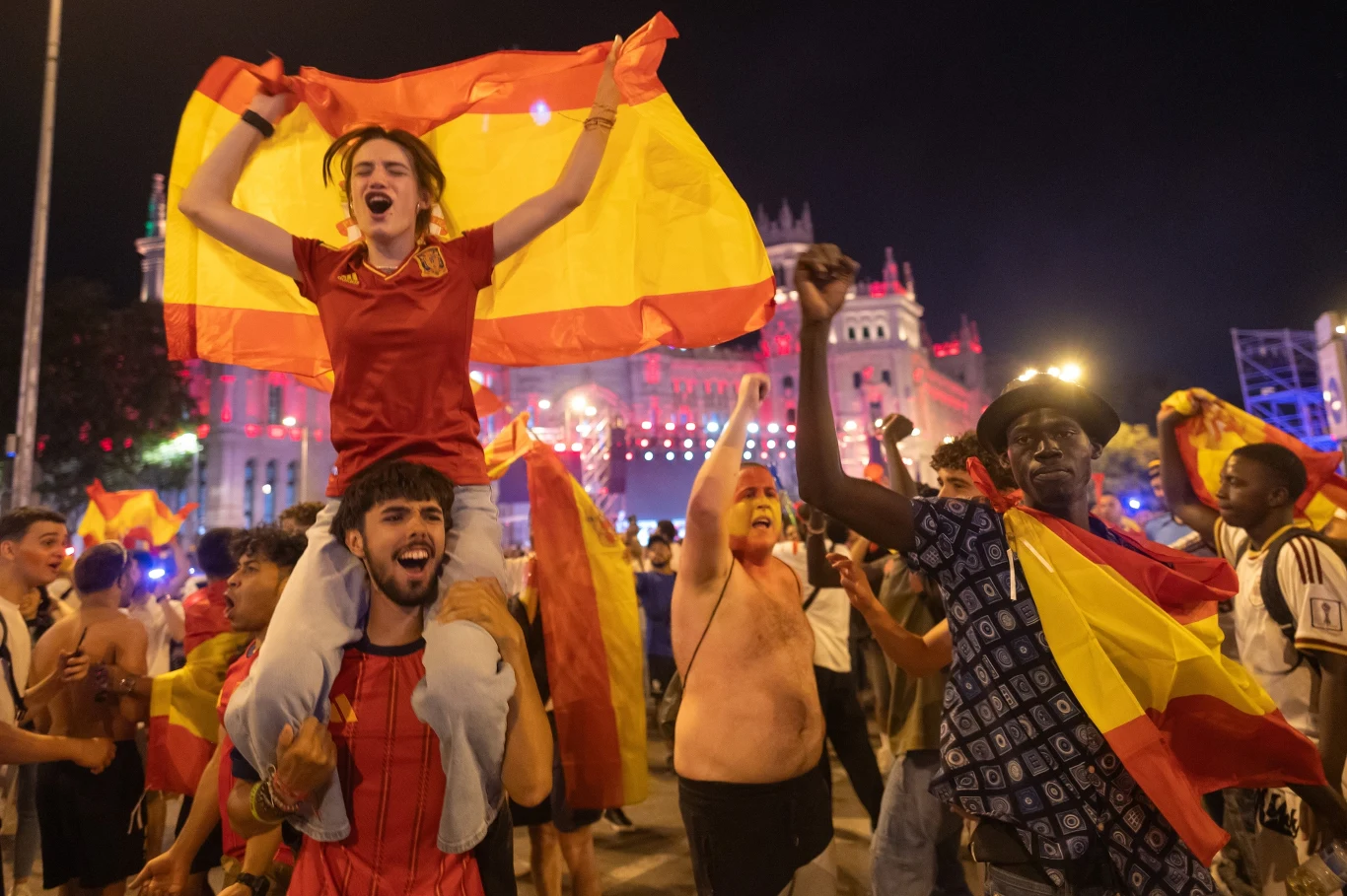 Tłum ludzi ubranych w barwy czerwono-żółte z flagami Hiszpanii świętuje na ulicy, jedna osoba siedzi na ramionach drugiej, unosi flagę i krzyczy z radości.