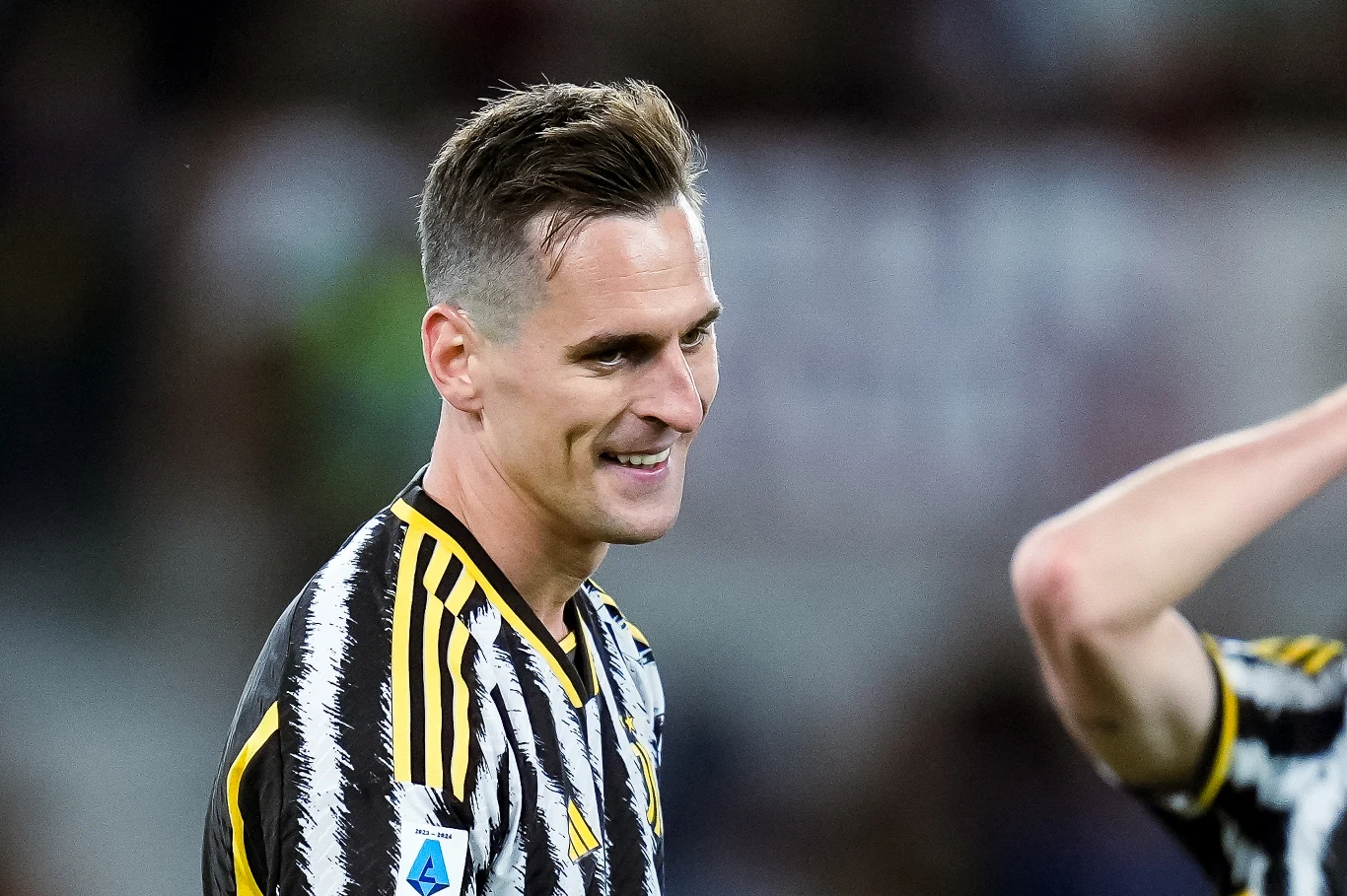 Piłkarz w pasiastym stroju sportowym z logo Serie A na rękawie uśmiecha się na boisku.