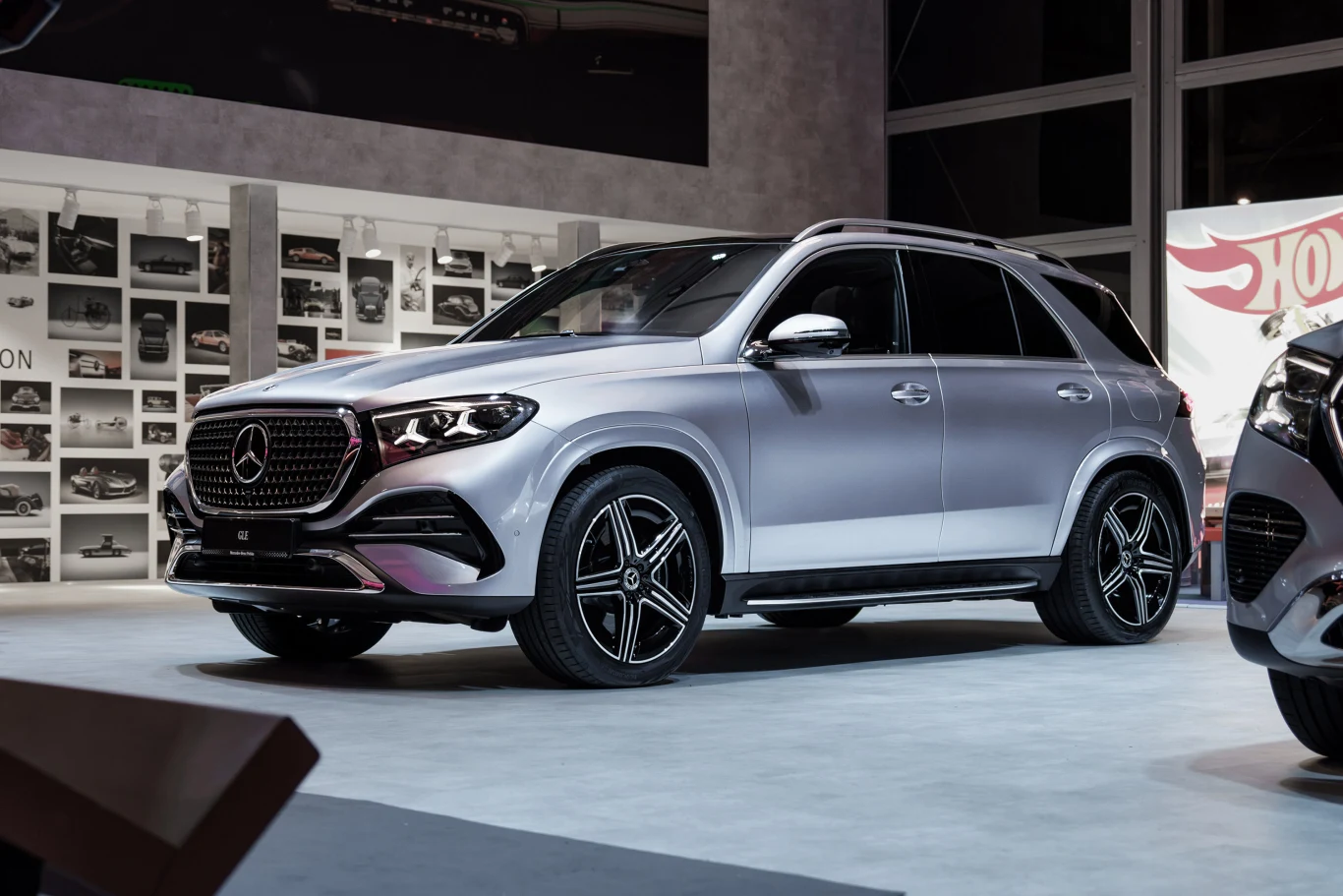 Srebrny SUV marki Mercedes-Benz z charakterystycznym grillem, czarnymi felgami i przyciemnianymi szybami, ustawiony na ekspozycji w otoczeniu nowoczesnego wnętrza.