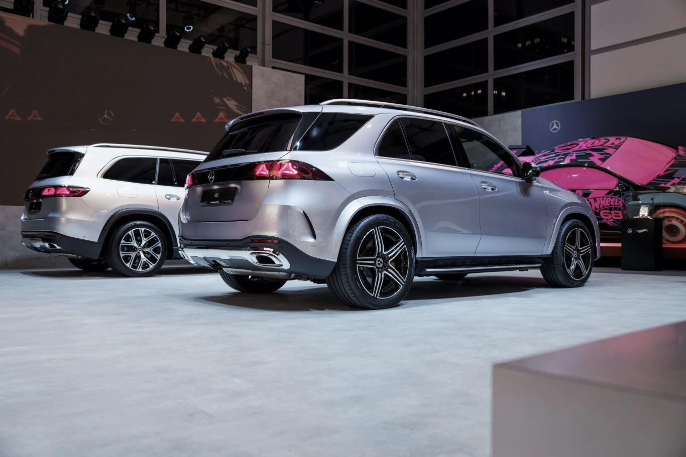 Dwa srebrne SUV-y marki Mercedes zaparkowane na ekspozycji, jeden z widocznym nowoczesnym tylnym zderzakiem i czarnymi felgami, w tle ściana z logotypem producenta.