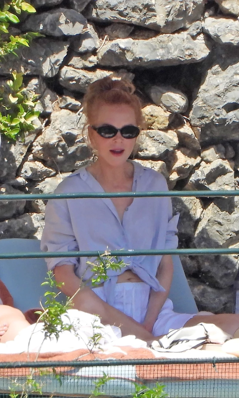 Nicole Kidman na wakacjach w Portofino Kobieta w jasnej koszuli i ciemnych okularach przeciwsłonecznych siedzi na leżaku przy kamiennym murze.